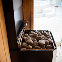 A sauna heater filled with smooth, rounded stones in a wooden room, conveying a warm and relaxing atmosphere. Light filters softly in the background.