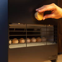 A hand adjusts a wooden knob on a dark, modern appliance with a grid, showing several round, light-colored objects inside. The scene is warmly lit.