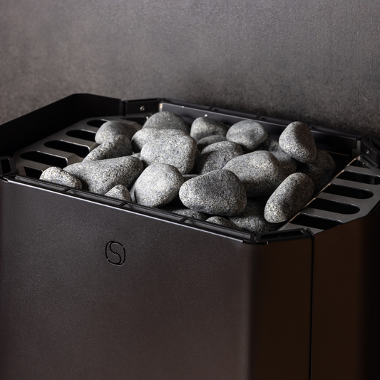A sauna heater filled with smooth gray stones against a dark wall. The metal grate supports the stones, creating a warm, tranquil atmosphere.