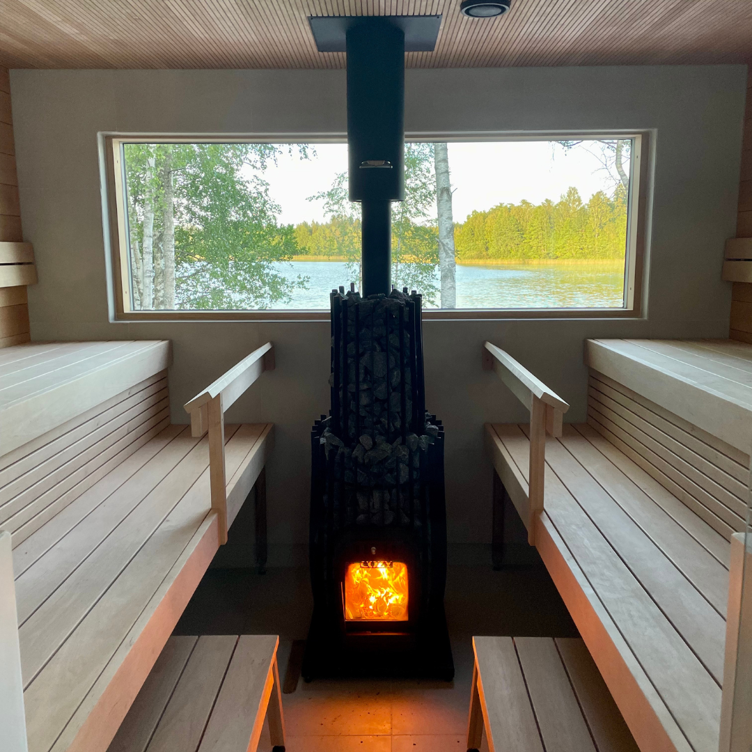 Sauna interior with wooden benches and a lit stove, featuring a window view of a lake and trees.