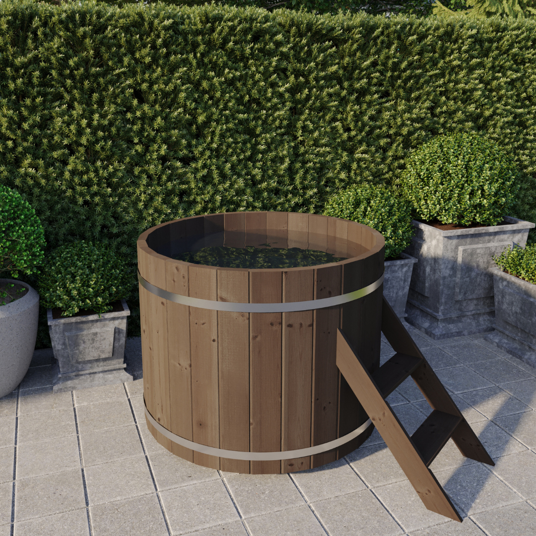 A round, brown cold tub made of wood, with a stainless steel ladder, placed on a tiled area outside with shrubbery in the background.