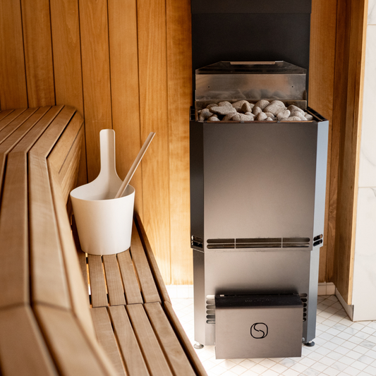 A sauna interior with wooden walls and bench. A modern sauna heater with stones is on the right, and a white water bucket with a ladle sits on the bench.