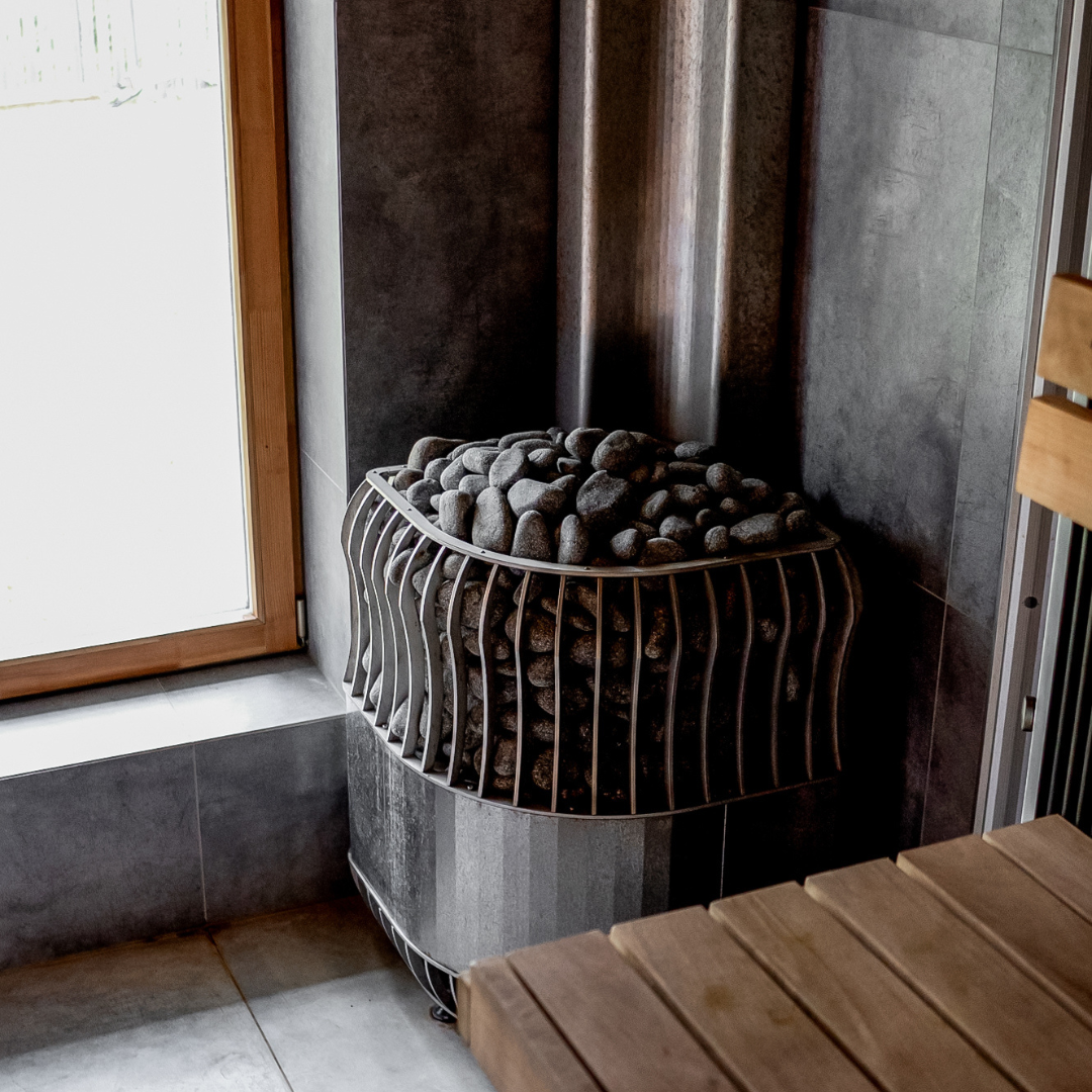 Modern sauna corner featuring a metal heater filled with smooth stones, wooden benches, and a large window. The atmosphere is peaceful and minimalist.