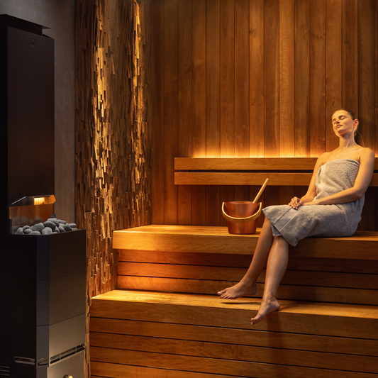 A woman in a towel relaxes on a wooden bench inside a warmly lit sauna room. A copper bucket and ladle rest nearby, exuding a serene ambiance.