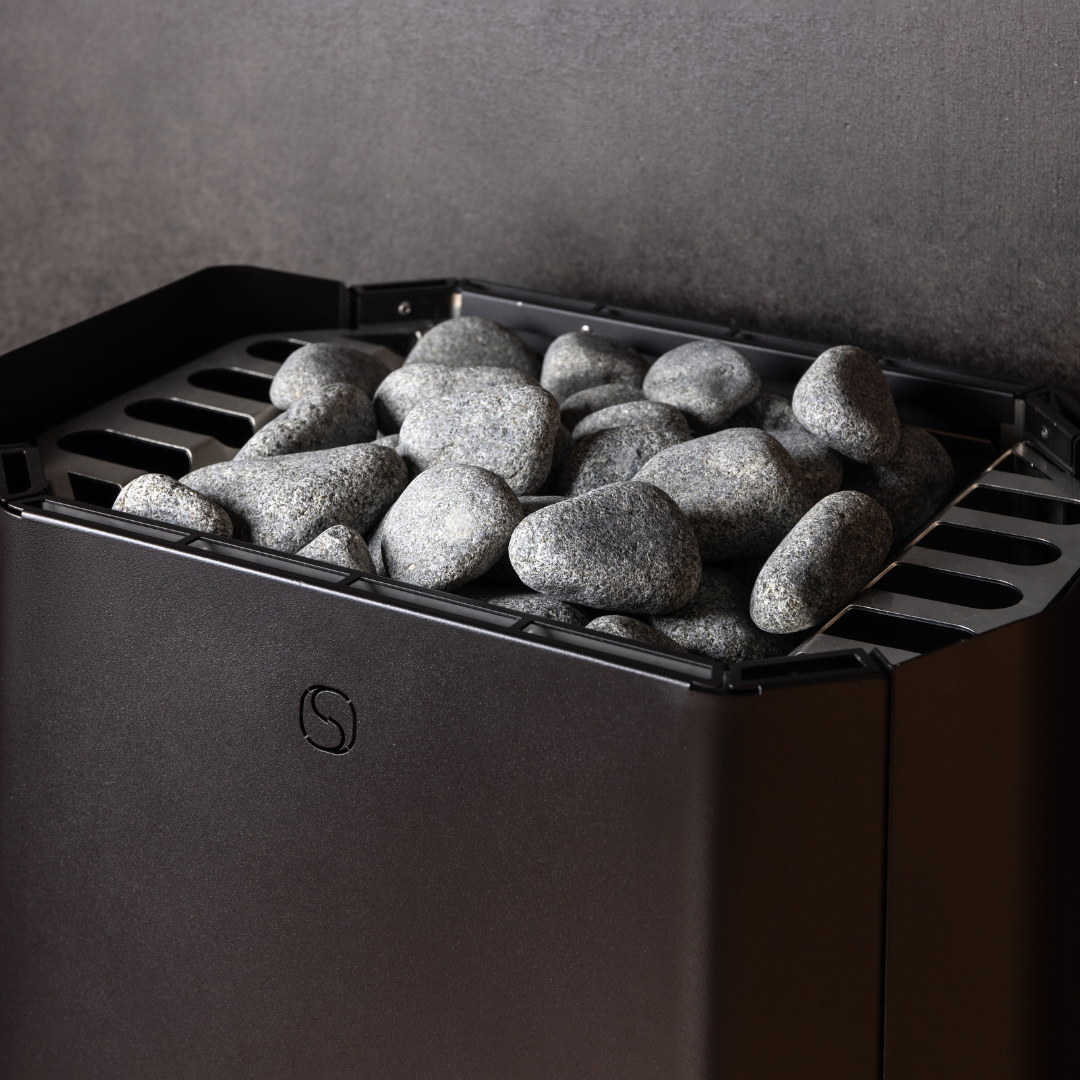 A sauna heater filled with smooth gray stones against a dark wall. The metal grate supports the stones, creating a warm, tranquil atmosphere.