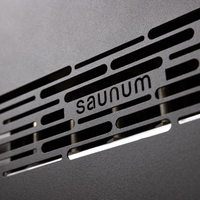 Close-up of a black metal surface with horizontal slits, showing the word "saunum" cut into the design. The tone is sleek and modern.