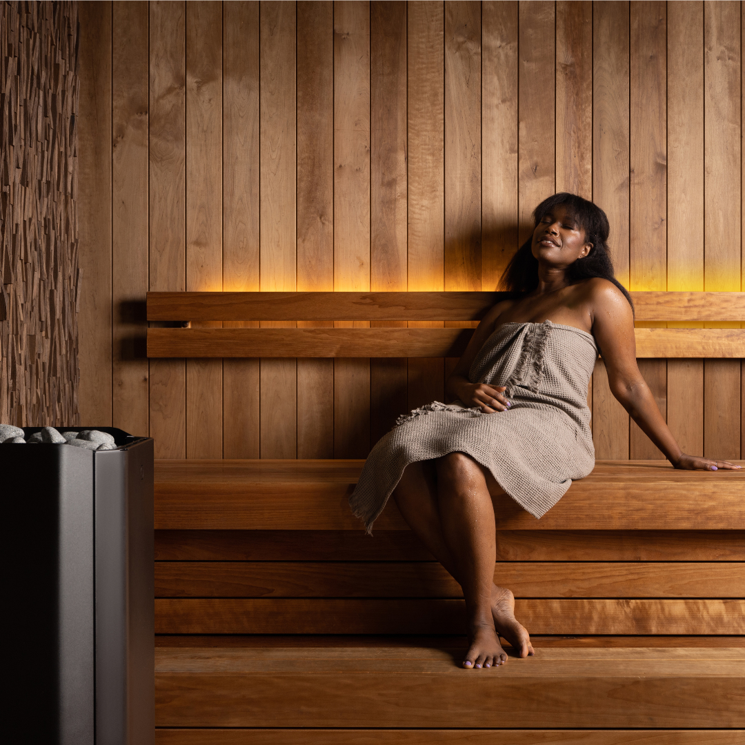 A woman relaxes on a wooden sauna bench, wrapped in a towel. The warm lighting and wooden panels create a serene and calming atmosphere.