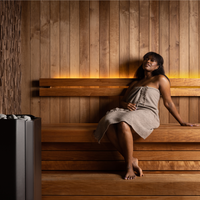 A woman relaxes on a wooden sauna bench, wrapped in a towel. The warm lighting and wooden panels create a serene and calming atmosphere.