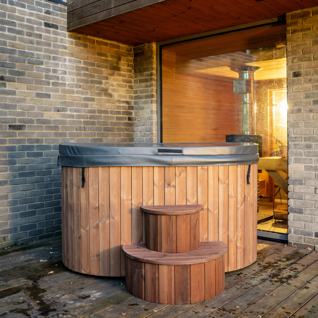 An outdoor wooden ice bath tub with ergonomic seats, situated on a deck with a scenic view of nature in the background. Includes insulated cover and wooden steps.