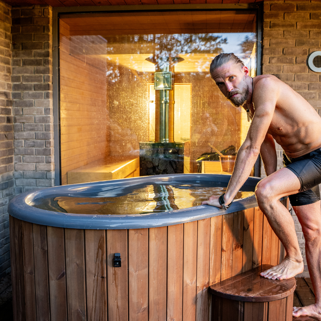 Man stepping into a wooden hot tub outside a sauna with large window. He's shirtless, wearing swim trunks, and the ambiance is warm and relaxing.