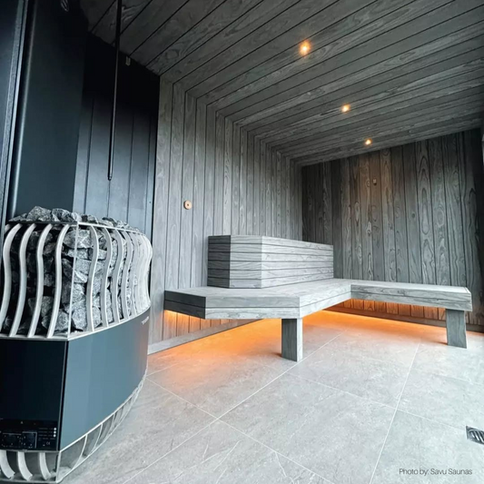 Modern sauna interior with gray wooden walls and benches. A black heater with sauna stones is on the left. Warm lights create a cozy ambiance.