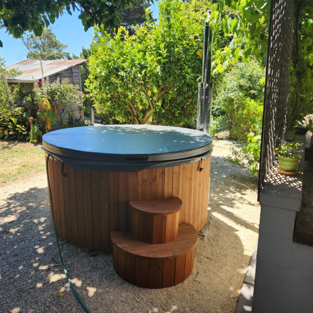 A covered wooden hot tub with small steps is set in a lush garden with green foliage and scattered sunlight, creating a serene, inviting atmosphere.